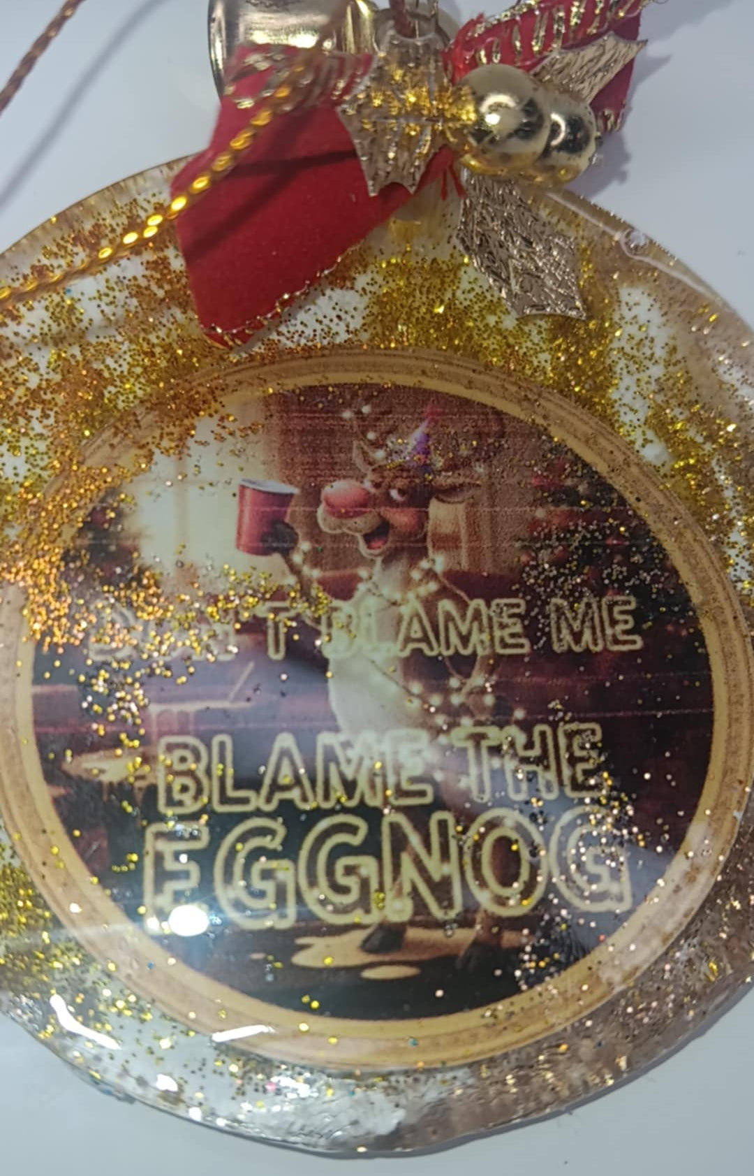 Decorative Christmas ornament with a reindeer and text 'Blame the Eggnog' on a gold and red background.